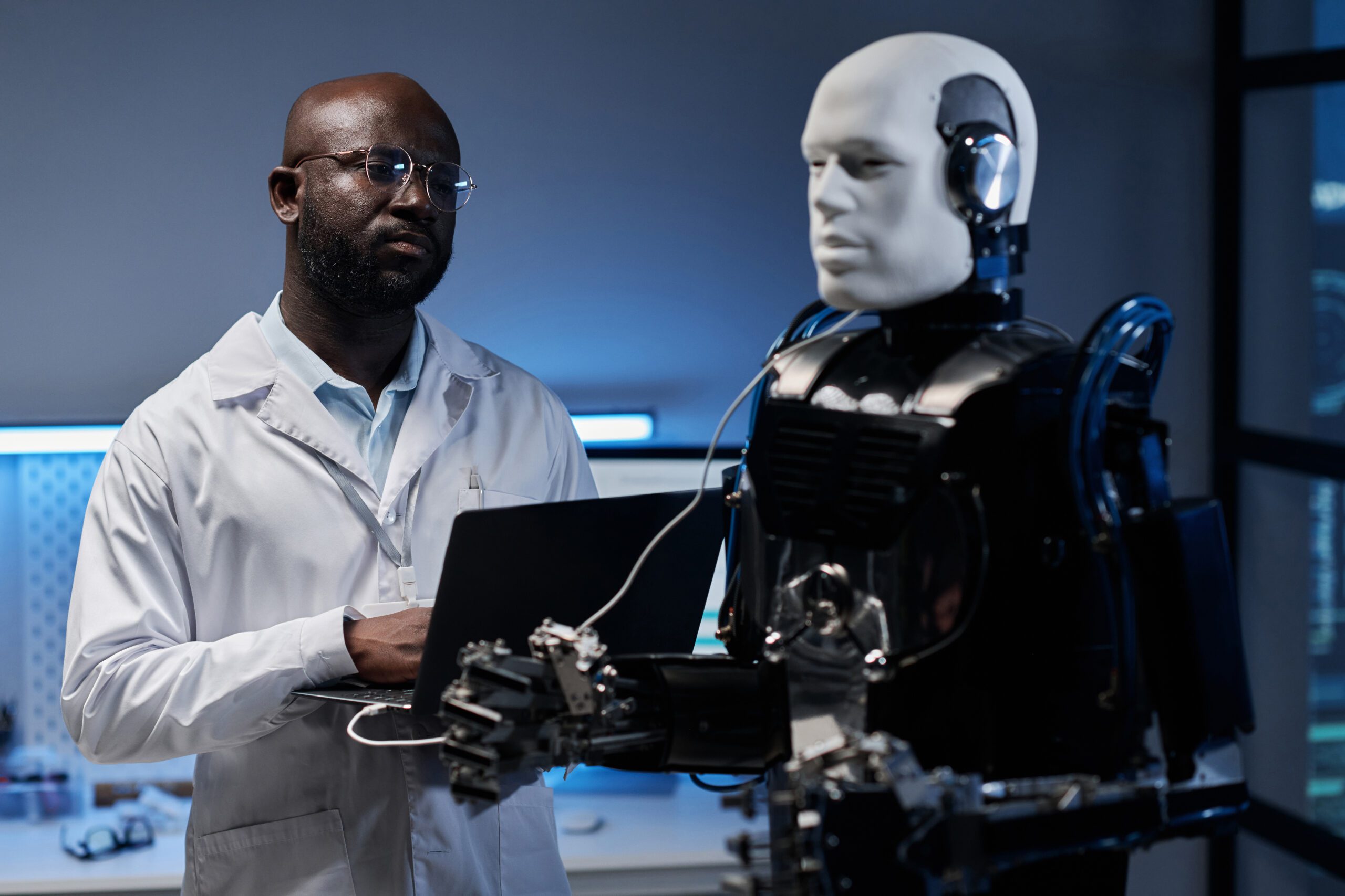African American engineer in lab coat using laptop to control the robot in office