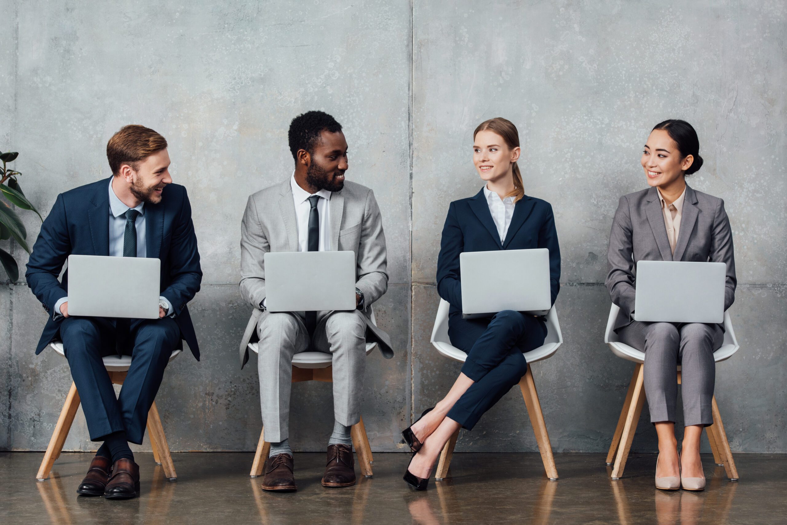 smiling multiethnic businesspeople sitting on chairs and using laptops in waiting hall