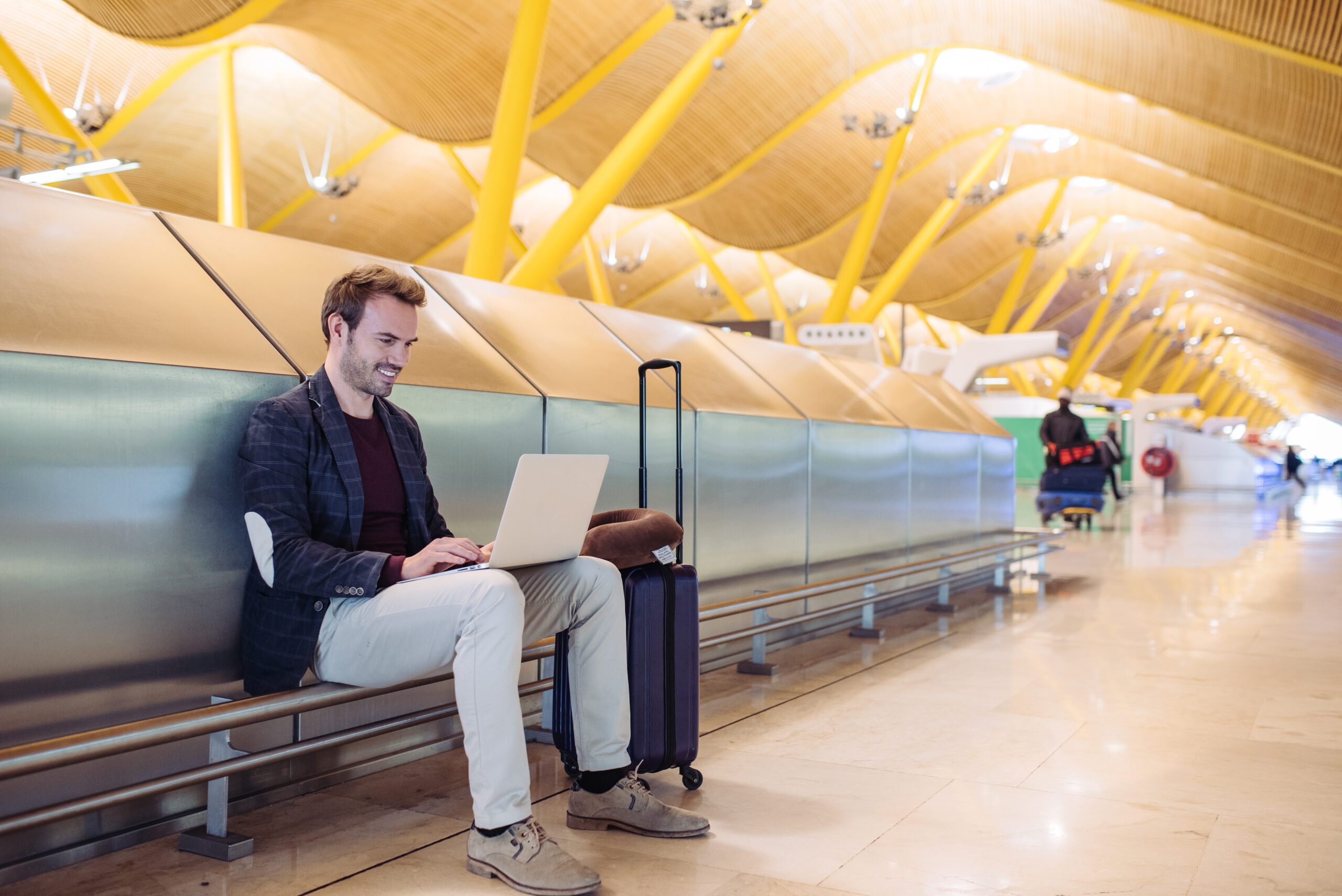Young attractive man sitting at the airport working in his laptop waiting his flight with a suitcase.